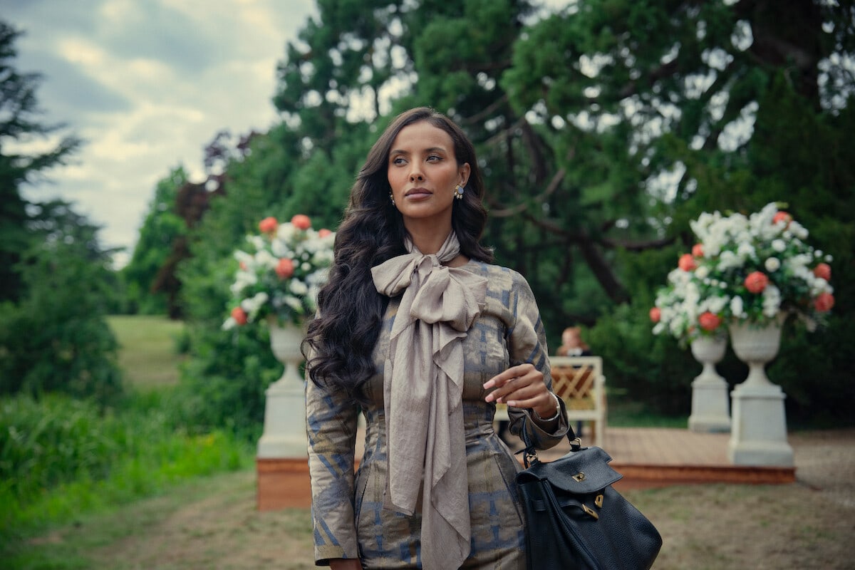 Aisha — Woman with long dark hair in an elegant outfit and bow scarf stands outdoors in a lush garden with large floral arrangements and greenery, holding a black handbag on her arm.