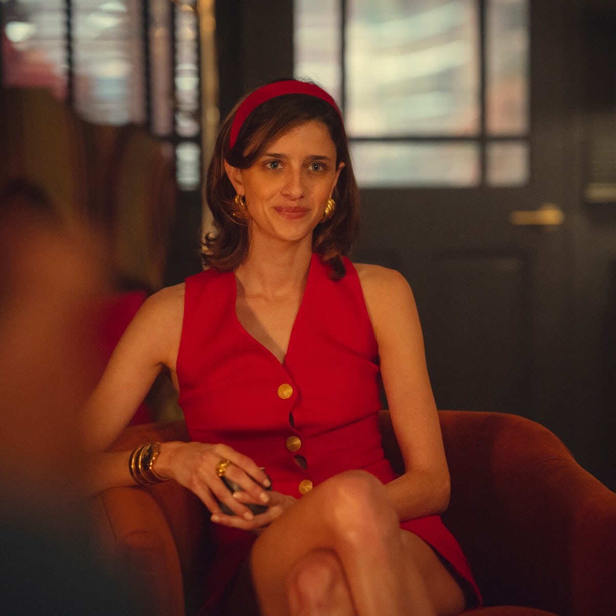 Bella — Woman in a sleeveless red dress with gold buttons and matching headband sits on an orange chair in a warmly lit, stylish indoor setting with blurred background and glass doors.