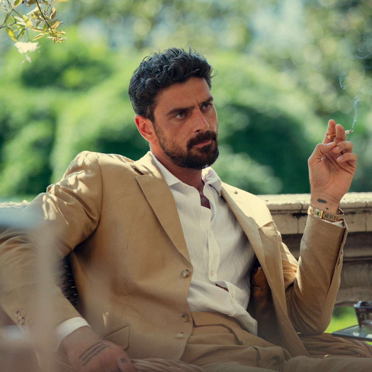 Cico Maldini — Man in a tan suit sitting outdoors on a patio, holding a cigarette, looking serious with greenery and sunlight in the background.