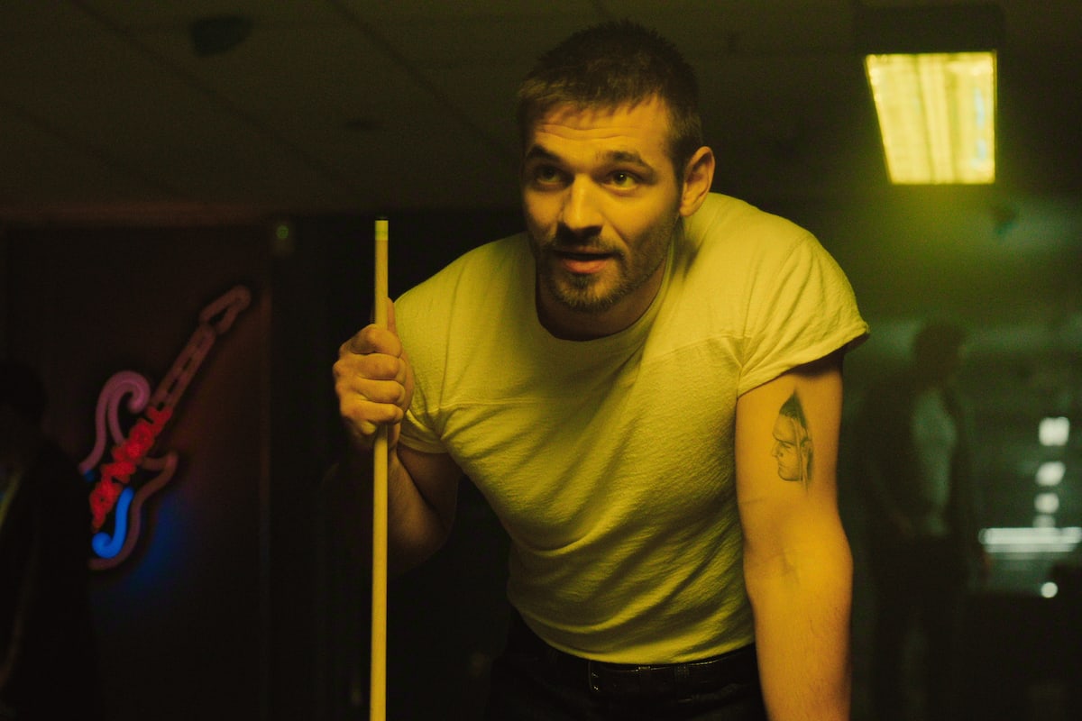 Joe Green — Man in white t-shirt holding pool cue, leaning over a pool table in a dimly lit room with neon guitar sign and overhead lights, focused and ready to play.