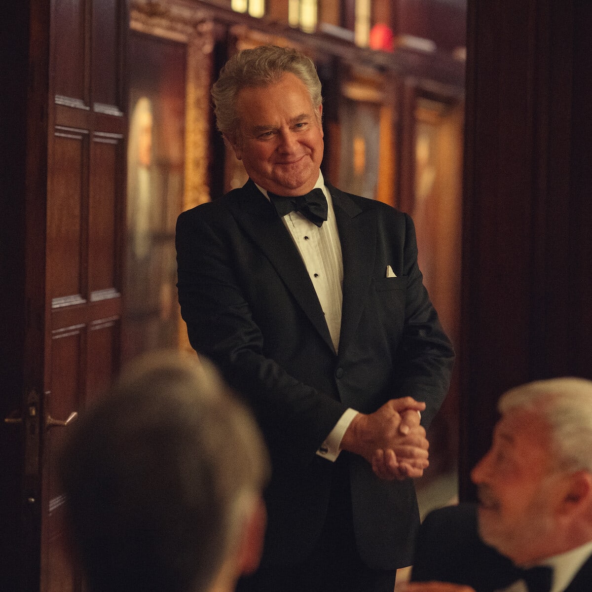 Lord Hawthorne — Man in a tuxedo smiling and standing in an elegant, wood-paneled room with portraits, while others in formalwear sit and interact in the foreground.