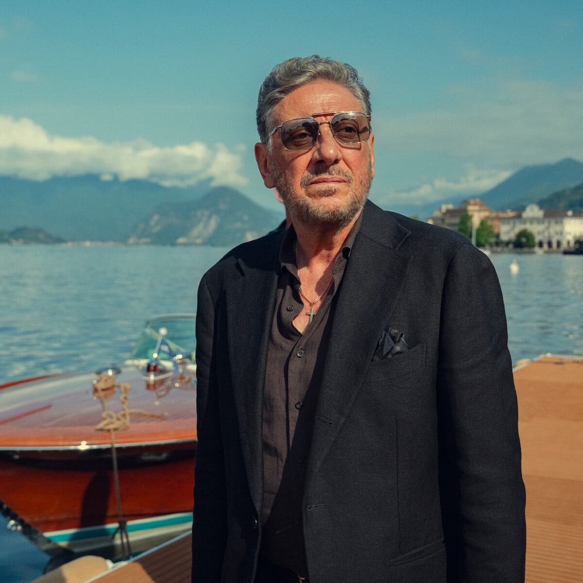 Marco Moretti — Man in a dark suit and sunglasses stands by a wooden boat on a dock, with a lake, mountains, and town buildings in the background under a clear sky.