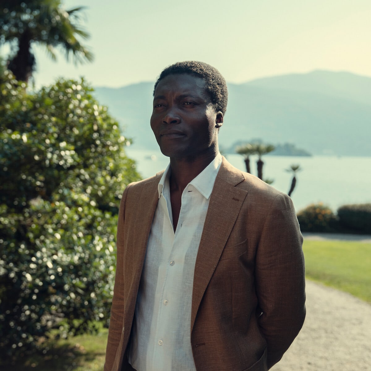 Nanny Asante — Man in a light brown suit and white shirt standing outdoors on a sunny day with greenery, palm trees, mountain lake, and distant hills in the background.